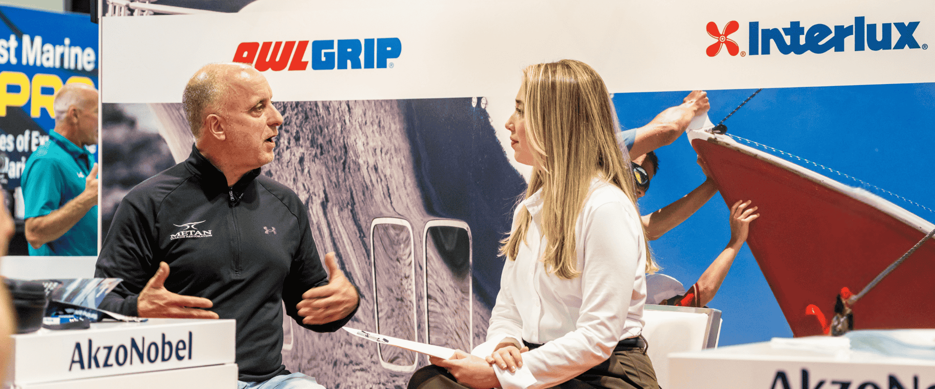 Mike Borrelli of Metan Marine speaking with a representative from AkzoNobel Yacht Coatings at the IBEX 2025 show, with Awlgrip and Interlux branding visible in the background.