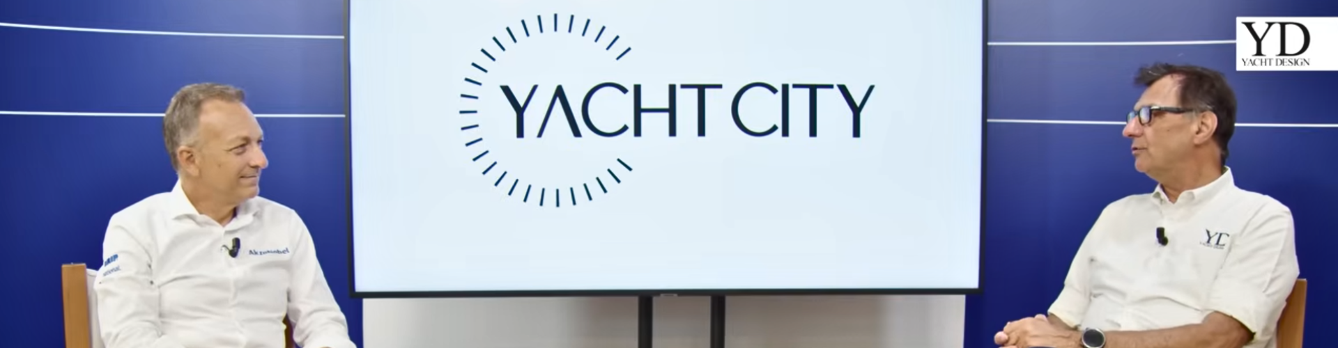 Two men seated and speaking in a studio interview setting, with a screen behind them displaying “Yacht City.”