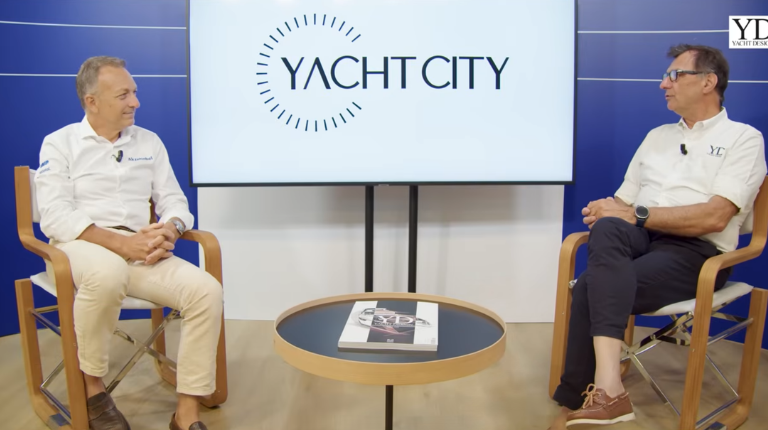 Two men seated and speaking in a studio interview setting, with a screen behind them displaying “Yacht City.”